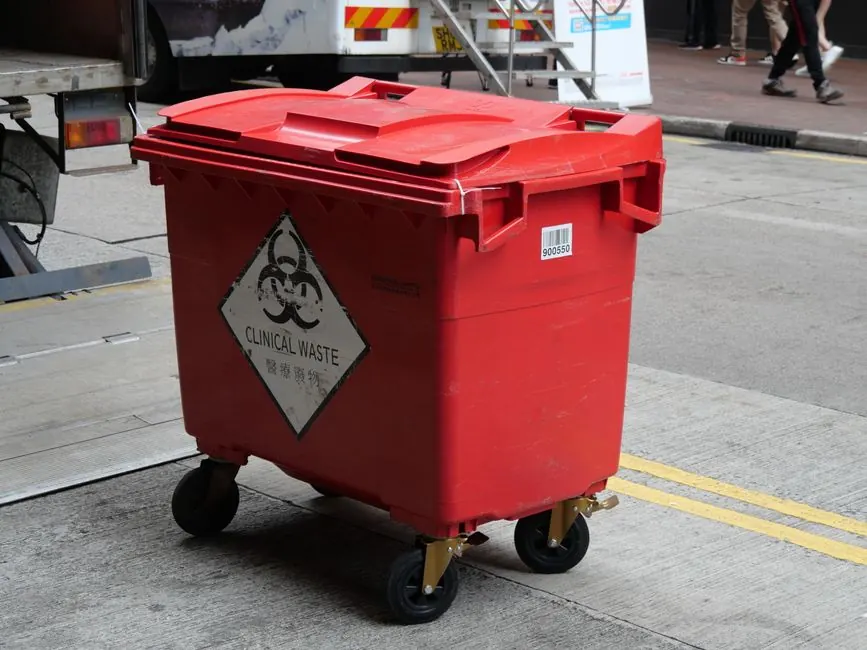 A red clinical waste bin labeled with biohazard symbol on a city street indicating proper waste management.
