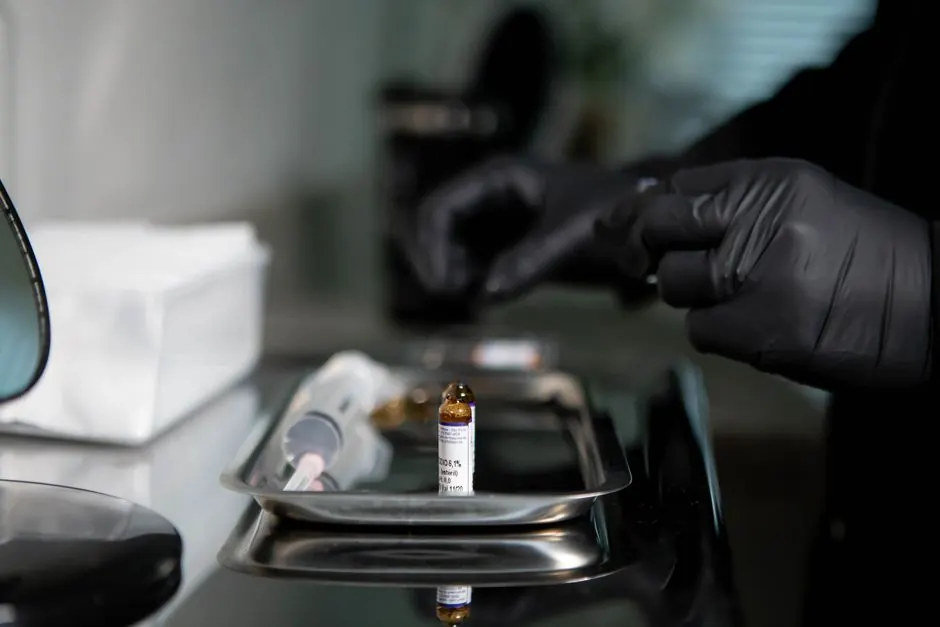 Close-up of gloved hands preparing a syringe with a vial on a metal tray in a clinical setting.